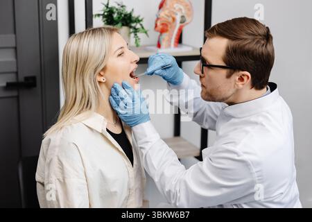 Il medico utilizza la spatola per ispezione per esaminare la gola del paziente. Medico ENT che fa esame di gola di una donna. La paziente aprì il check-up bocca-gola. Foto Stock