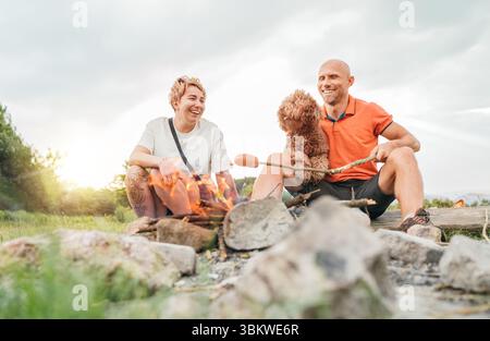 Graziosa scena di picnic all'aperto: Coppia felice sorridente con il cane Maltipoo che grigia salsicce su bastoncini da barbecue seduti accanto al fuoco durante il tour di trekking Foto Stock