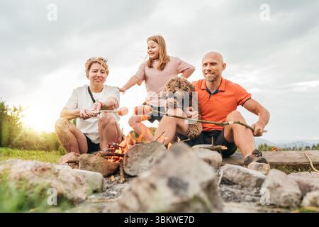 Graziosa scena di picnic all'aperto: Padre, figlia e mamma con il cane Maltipoo che grigliava salsicce su bastoncini da barbecue seduti accanto al fuoco durante il tour escursionistico Foto Stock