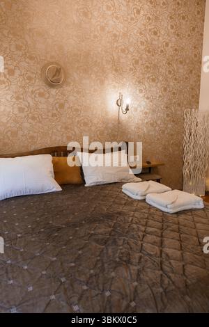 Una camera da letto illuminata e' caratterizzata da un letto marrone ben fatto con cuscini bianchi e asciugamani piegati, su uno sfondo di carta da parati beige e pareti in stile rustico Foto Stock
