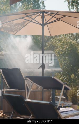 Un tranquillo giardino estivo sul retro con un ombrello beige da patio, una griglia con lieve fumo, due poltrone e un sistema di appannamento creano un "i" Foto Stock