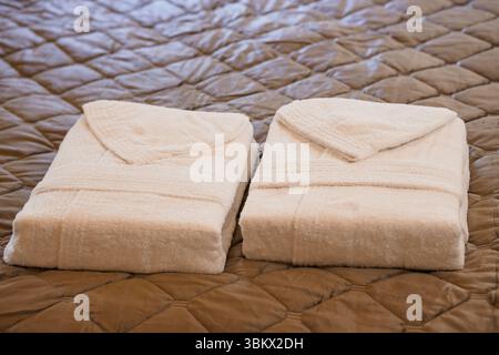 Due accappatoi piegati color crema si trovano ordinatamente su un copriletto trapuntato marrone che evoca un senso di lusso, comfort spa e relax in un ambiente alberghiero Foto Stock