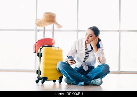 Giovane donna in attesa in aeroporto, seduta con valigia e oggetti essenziali, con il telefono, che viaggia per una destinazione di vacanza Foto Stock