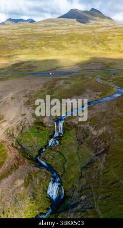 Una vista mozzafiato della cascata di Selvallafoss che scorre in un fiume sereno, circondato da un terreno verde lussureggiante e dalle montagne spettacolari nelle terre ghiacciate, la splendida natura selvaggia durante la luce del mattino presto. Foto Stock