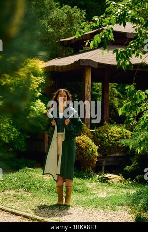 Una giovane donna con un impermeabile alla moda si erge orgogliosamente alla luce del sole tra una vegetazione lussureggiante. Foto Stock