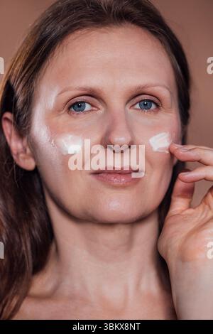 Una donna di mezza età applica delicatamente la crema idratante sulla guancia. Una signora di mezza età si strofina il viso con una crema anti-invecchiamento. Ritratto ravvicinato di un wom Foto Stock