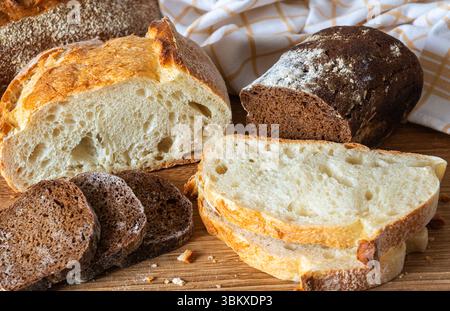 Diversi tipi di pane appena sfornato disposti su un tagliere di legno, che mostra le loro consistenze e colori con un panno nelle vicinanze. Foto Stock