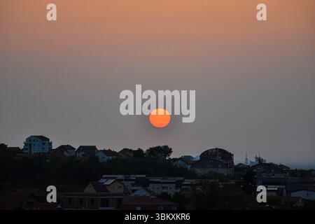 Il sole dorato scintillante tramonta dietro il paesaggio urbano, con il cielo limpido che sbiadisce dall'arancione profondo al blu fino al grigio bluastro tenue Foto Stock