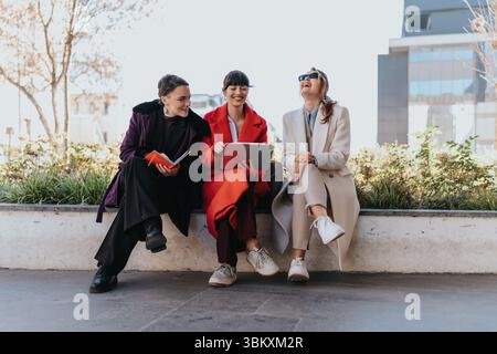 Tre donne d'affari che collaborano e ridono mentre sono sedute all'aperto in un ambiente urbano moderno Foto Stock