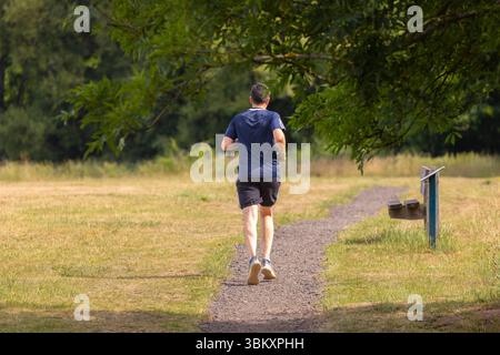 Vista posteriore di un uomo isolato di mezza età con pantaloncini corti e T-shirt che corre in un parco del Regno Unito in una giornata di sole. Foto Stock