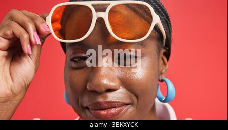Donna nera, ritratto e occhiali da sole con trucco in studio per la moda su sfondo rosso. Faccia, persona femminile o modello africano con sorriso o bastoncini Foto Stock