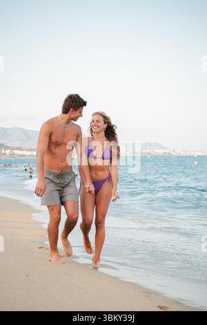 Una coppia allegra passeggia mano nella mano lungo la splendida spiaggia di sabbia, godendosi il caldo sole e la dolce brezza dell'oceano mentre crei un'atmosfera molto apprezzata Foto Stock