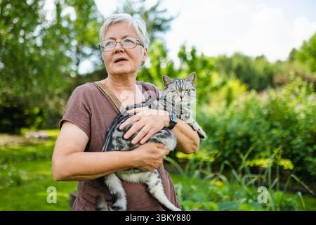 Una donna anziana tiene delicatamente un gatto Shorthair britannico in un giardino lussureggiante, entrambi guardando attentamente in lontananza in una giornata calda. Foto Stock