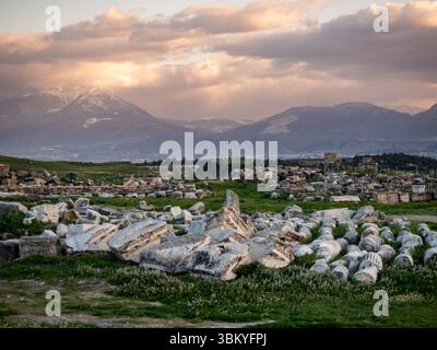 L'immagine mostra le rovine di Hierapolis, un'antica città situata vicino a Pamukkale nell'odierna Turchia. Le rovine includono colonne cadute e altri frammenti architettonici, sparsi in un campo erboso. Sullo sfondo, le montagne possono essere viste sotto un cielo nuvoloso, aggiungendo alla bellezza paesaggistica del sito storico. Foto Stock
