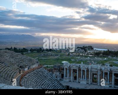 L'immagine mostra l'antica città di Hierapolis in Turchia, con le rovine del teatro in primo piano e le travertini Pamukkale sullo sfondo. Il sole sta tramontando, proiettando un caldo bagliore sul paesaggio. Foto Stock
