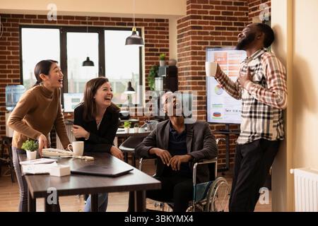 Un gruppo multirazziale di professionisti nell'ufficio murale si prende una pausa, chiacchierando e ridendo insieme. L'uomo afroamericano con tazza di caffè condivide una storia divertente con la sua squadra. Foto Stock