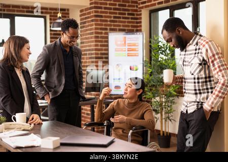 Team di startup multietnico in piedi e ad ascoltare la loro collega donna in sedia a rotelle mentre condivide una storia divertente. Diversi gruppi di dipendenti si uniscono durante la loro pausa in uno spazio di lavoro moderno. Foto Stock