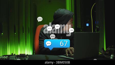 Uomo che digita indossando una felpa scura con cappuccio che preme i tasti del laptop nella sala server, con bolla di notifica 67 Foto Stock