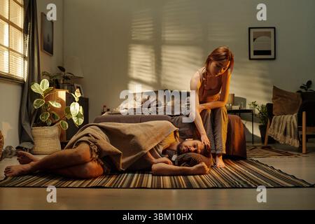 Donna che conforta il giovane uomo adulto sdraiato sul pavimento vicino al letto, donna che tocca delicatamente la testa di mans, entrambi mostrando segni di disagio emotivo in camera da letto Foto Stock