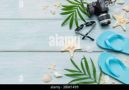 Infradito, macchina fotografica, occhiali da sole, conchiglie e foglie di palma che creano una sistemazione per le vacanze estive su uno sfondo di legno blu Foto Stock