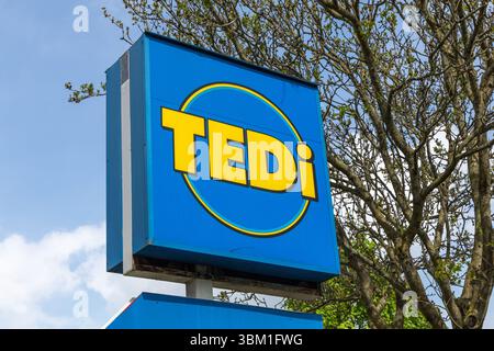 Un cartello blu luminoso del negozio Tedi con scritta gialla e un design circolare si staglia contro un cielo parzialmente nuvoloso. Kleve, Germania. 22 aprile 2025. Foto Stock