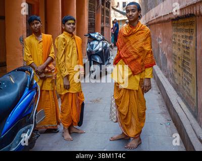 India, Varanasi, 16 marzo 2025. Tre giovani pellegrini in vesti di zafferano si stendono a piedi nudi in una stretta corsia vicino ai ghat, portando con sé borse di stoffa tradizionali Foto Stock