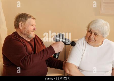 Uomo anziano che usa una pistola per massaggi per alleviare il dolore muscolare nelle donne anziane a casa, sorridendo e legando momento. Foto Stock