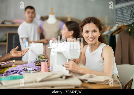 Donna di mezza età che cucisce mentre altre lavorano con macchina da cucire e manichino Foto Stock