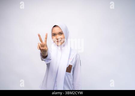 Eccitata studentessa indonesiana delle scuole superiori in uniforme bianca e grigia dando il numero due con gesti a mano Foto Stock