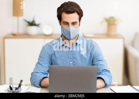 Uomo d'affari che indossa la maschera facciale che lavora sul laptop seduto sul luogo di lavoro Foto Stock