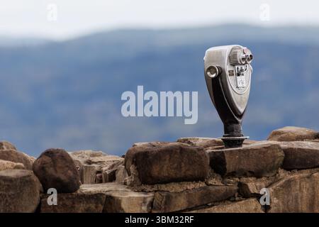 Visore binoculare a gettoni montato su una parete di pietra intemprata che si affaccia sulle lontane montagne blu sotto un cielo coperto in un ambiente panoramico all'aperto. Foto Stock