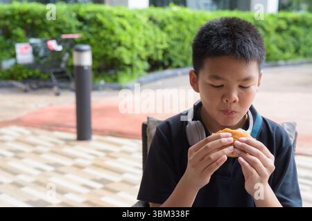 Studente thailandese che indossa le cuffie mangiando hamburger al tavolo all'aperto Foto Stock