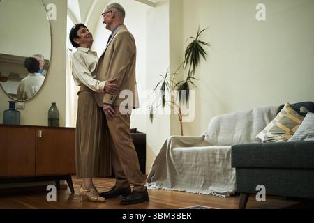 Coppie di anziani che ballano e si abbracciano in un soggiorno moderno con mobili e decorazioni contemporanee intorno. Grande specchio e pianta in vaso visibile sullo sfondo Foto Stock