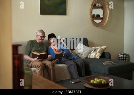 Una coppia di anziani seduta sul divano che legge libri e usa un tablet in salotto, creando un'atmosfera serena e intima che illustra la tecnologia che colma le lacune generazionali Foto Stock