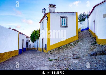 Stretta strada acciottolata nella medievale Obidos, Portogallo, fiancheggiata da tradizionali case imbiancate di bianco caratterizzate da colorate finiture gialle e blu. Foto Stock