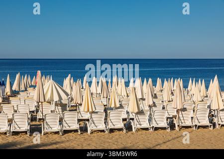 Una tranquilla mattinata d'estate su una spiaggia sabbiosa con file di ombrelloni chiusi e lettini bianchi di plastica di fronte al tranquillo Mar Mediterraneo sotto le nuvole Foto Stock