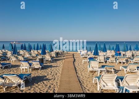 Una tranquilla mattinata d'estate su una spiaggia sabbiosa vicino al tranquillo Mar Mediterraneo con passerella in legno attraverso file di lettini bianchi vuoti e ombrello blu chiuso Foto Stock