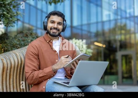 Un uomo indiano sorridente che indossa le cuffie lavora all'aperto con un notebook e un notebook. E' di fronte ad un edificio moderno. Foto Stock