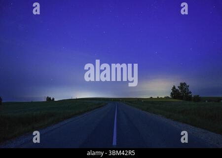 Una strada rurale deserta si estende all'orizzonte sotto un cielo stellato, illuminato da un debole crepuscolo e da un inquinamento luminoso lontano. Foto Stock