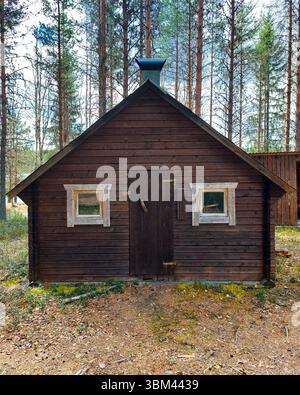 La cabina di legno scuro nel nord della Svezia si erge tranquillamente tra alti pini sotto un cielo parzialmente nuvoloso. Foto Stock