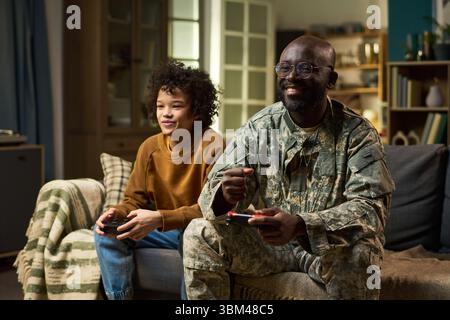 Uomo nero di mezza età in uniforme militare seduto sul divano a giocare con il bambino nero sorridente accanto a lui a casa, sia guardando lo schermo della televisione, momento di legame familiare Foto Stock