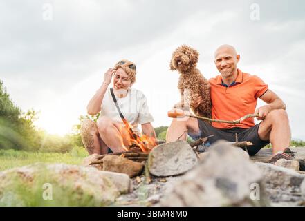 Graziosa scena di picnic all'aperto: Coppia felice sorridente con il cane Maltipoo che grigia salsicce su bastoncini da barbecue seduti accanto al fuoco durante il tour di trekking Foto Stock