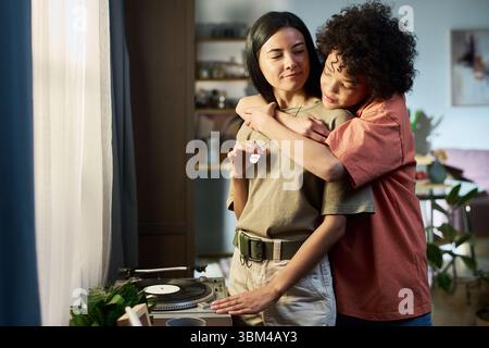 Donna caucasica di mezza età in piedi accanto a un giocatore di dischi che viene abbracciata da un adolescente nero a casa, sorridente e godendo del tempo in famiglia in salotto Foto Stock