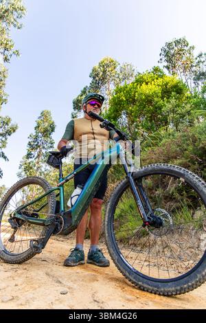 Uomo anziano che indossa abbigliamento da ciclismo e casco in piedi accanto alla sua mountain bike elettrica su un sentiero nella foresta, facendo una pausa durante un'escursione in na Foto Stock