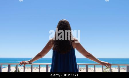 Giovane donna su un balcone che gode di una vista soleggiata sulla spiaggia in vacanza Foto Stock