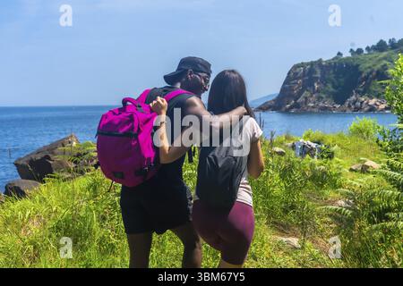 Coppia interrazziale con zaini che camminano su un sentiero vicino al mare, abbracciando e godendo la vista panoramica durante una soleggiata giornata estiva Foto Stock