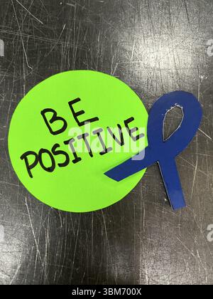 Un cerchio verde con un nastro blu e le parole devono essere positive. Il cerchio è su una superficie nera. Concetto di cancro Foto Stock