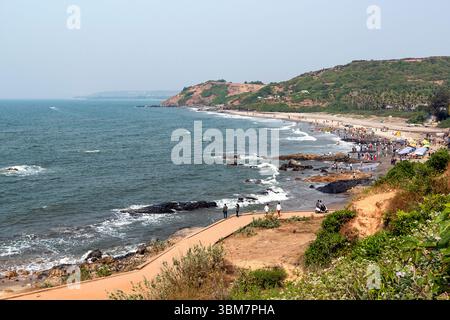 A Anjuna Beach a Goa, India, le onde dolci si infrangono su aspre formazioni rocciose lungo la costa, mentre numerosi visitatori si impegnano in attività sulla spiaggia Foto Stock