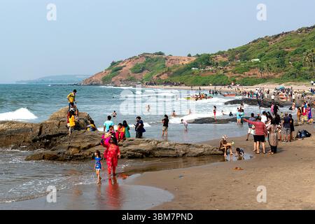 A Anjuna Beach a Goa, India, le onde dolci si infrangono su aspre formazioni rocciose lungo la costa, mentre numerosi visitatori si impegnano in attività sulla spiaggia Foto Stock