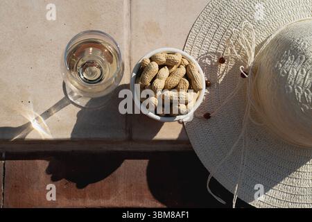 Un piatto dall'alto che offre gli indispensabili per le vacanze estive: Un bicchiere di vino bianco, una ciotola di arachidi e un cappello di paglia, con forti ombre grafiche Foto Stock